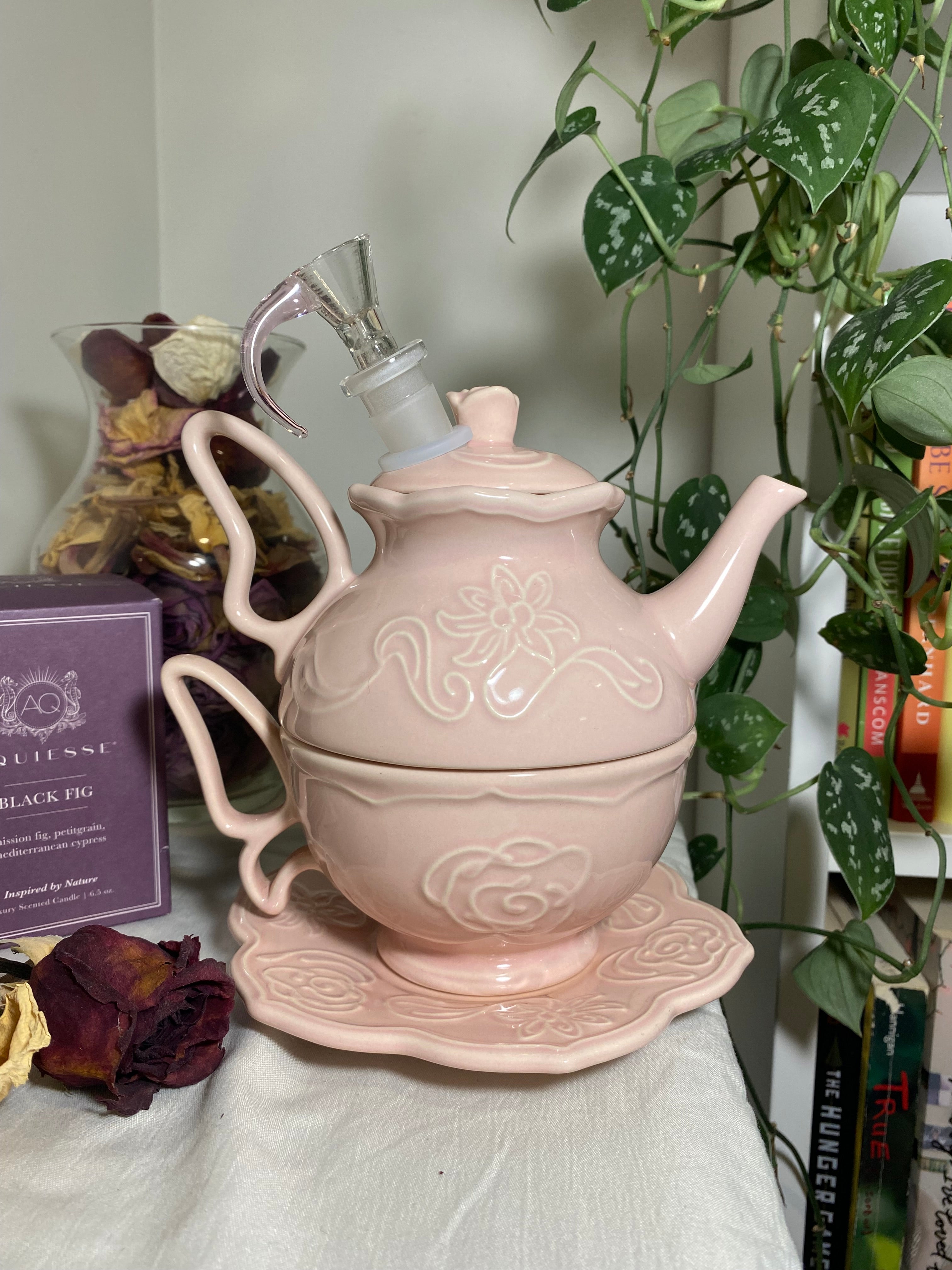A stackable light pink tea set for one that has been turned into a bong, cup, and saucer set. It’s being staged on a bookshelf with a plant and dried flowers in the background. 
